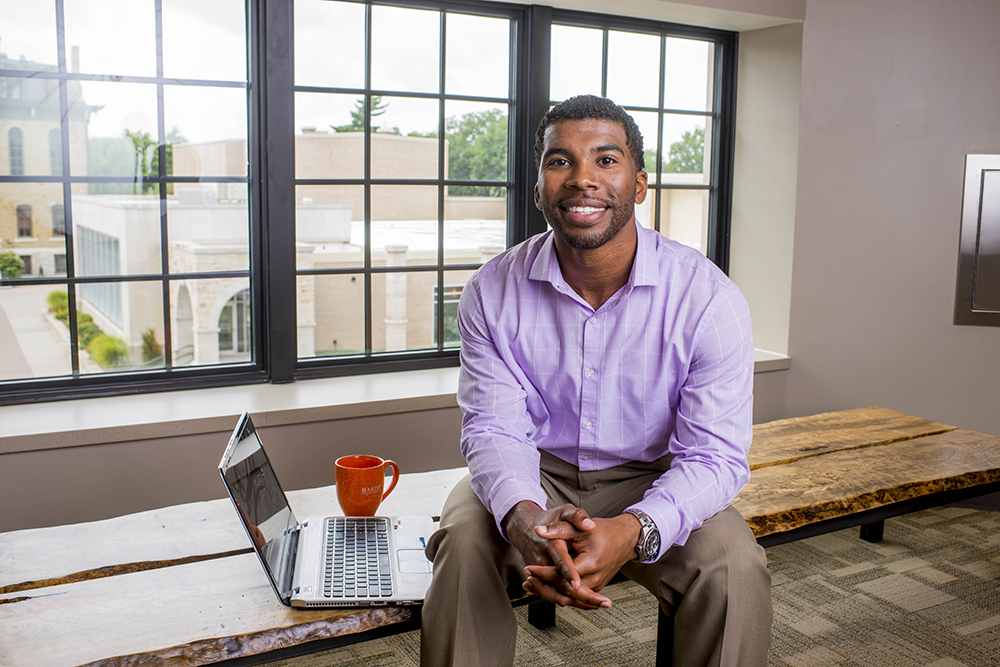 Student smiling, sitting on a table with a laptop and coffee mug next to him