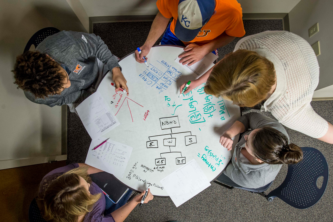 Overhead photo of five business analytics students working on a whiteboard table for a class project