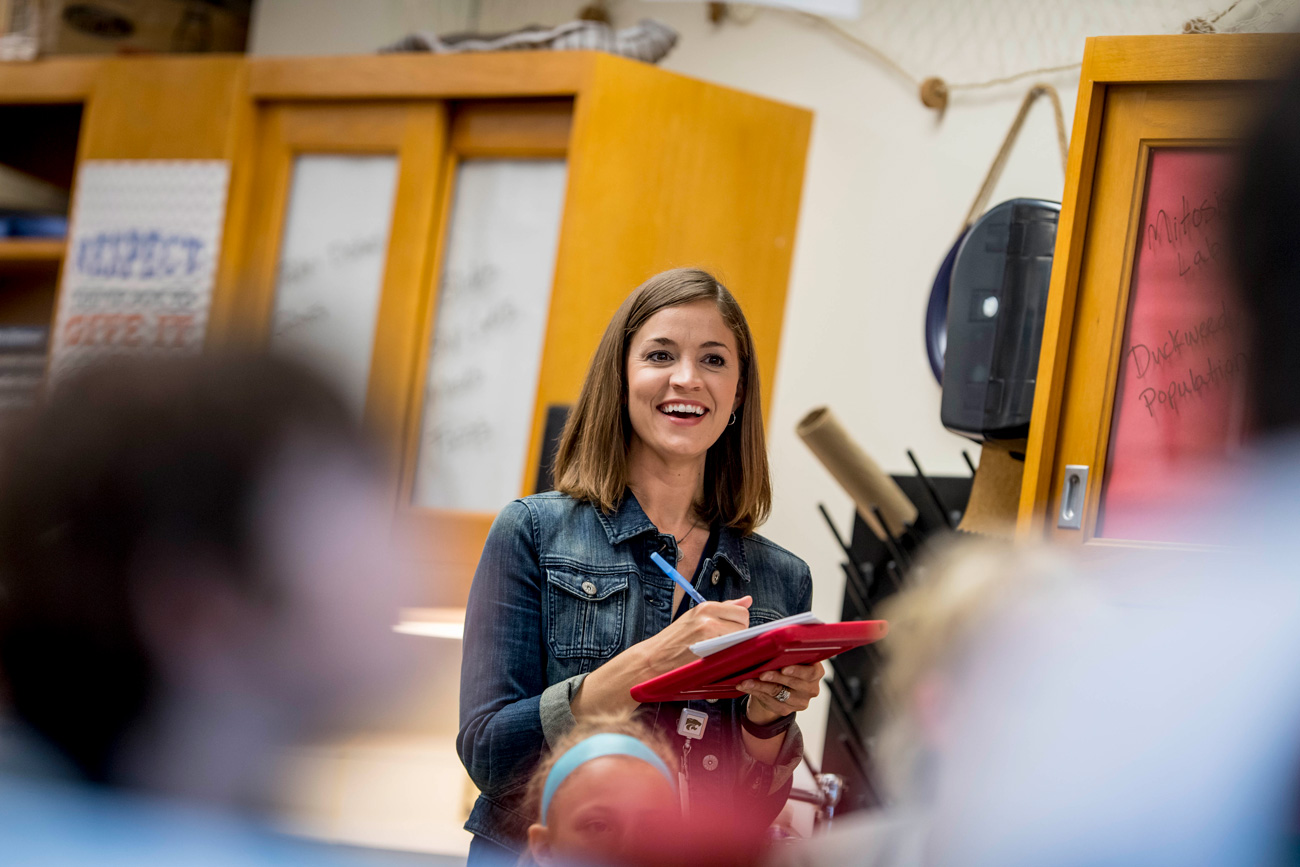 educator smiling using a tablet in a classroom with students