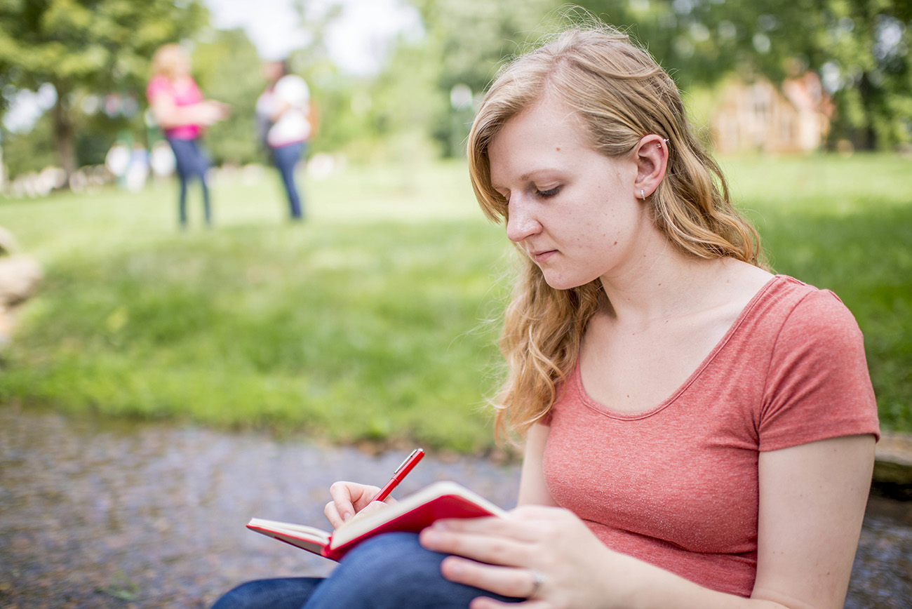 Student sitting outside on campus writing in a notebook for an english class