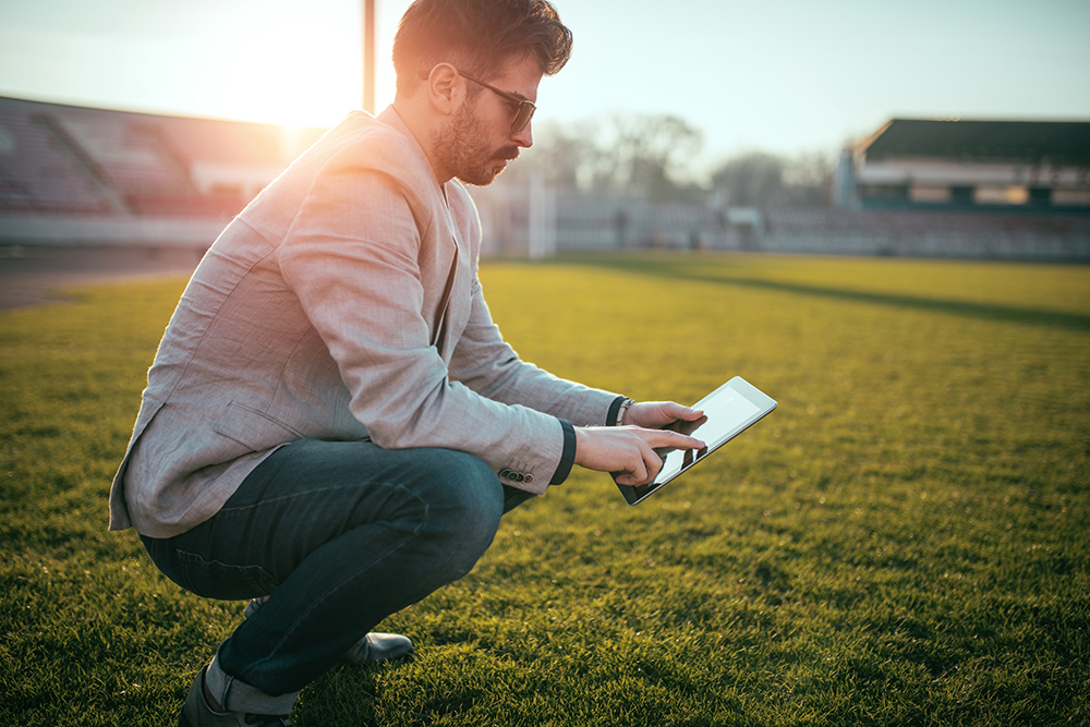 Student squatting in the grass with a tablet on the field