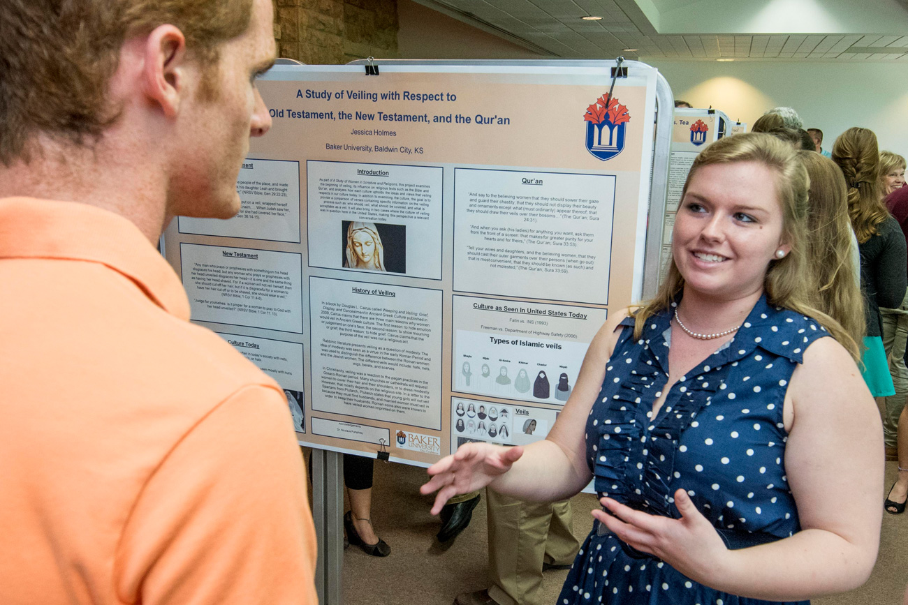 Religious Studies student standing in front of a research poster smiling and explaining her research to another student during dialogos presentation