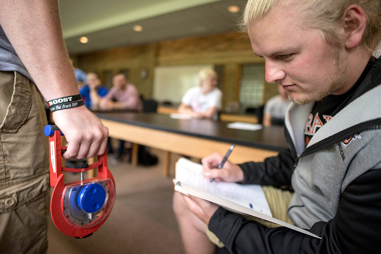 student taking notes in a sports administration classroom, observing another student holding a tool