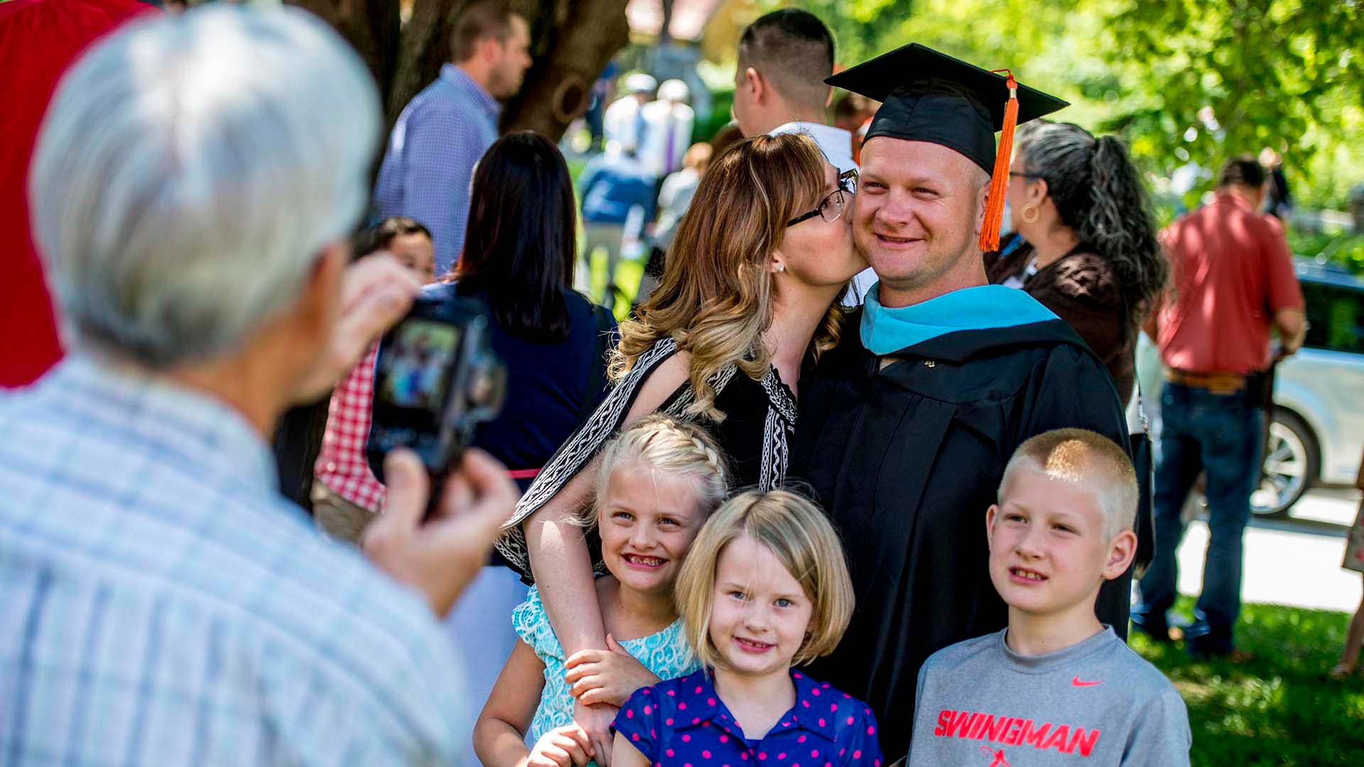 Master's graduate in a cap and gown receiving a kiss on the cheek with three kids in front of him, all posing for a photo