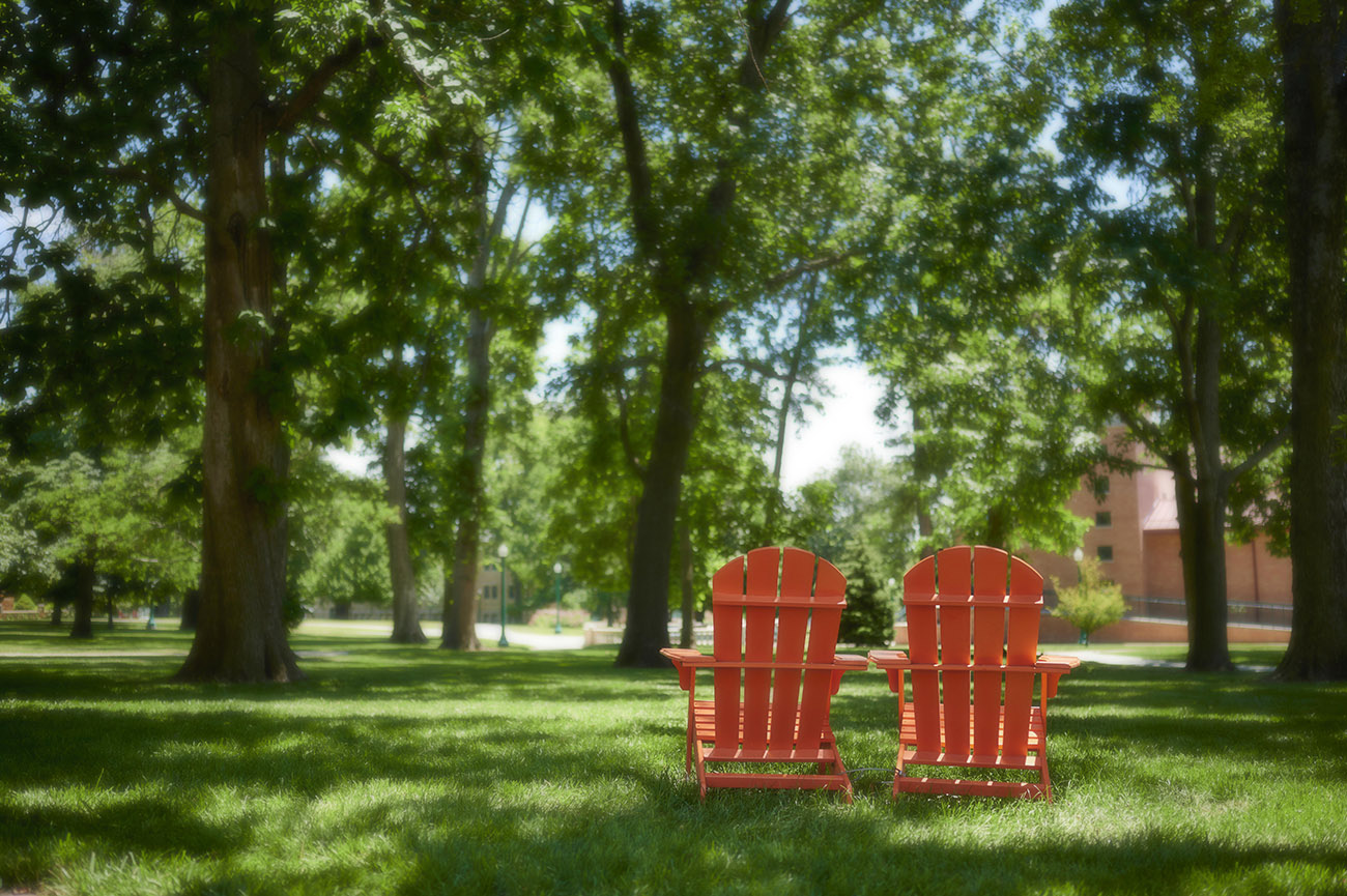 Two orange lawn chairs outside on campus in the grass, under the trees