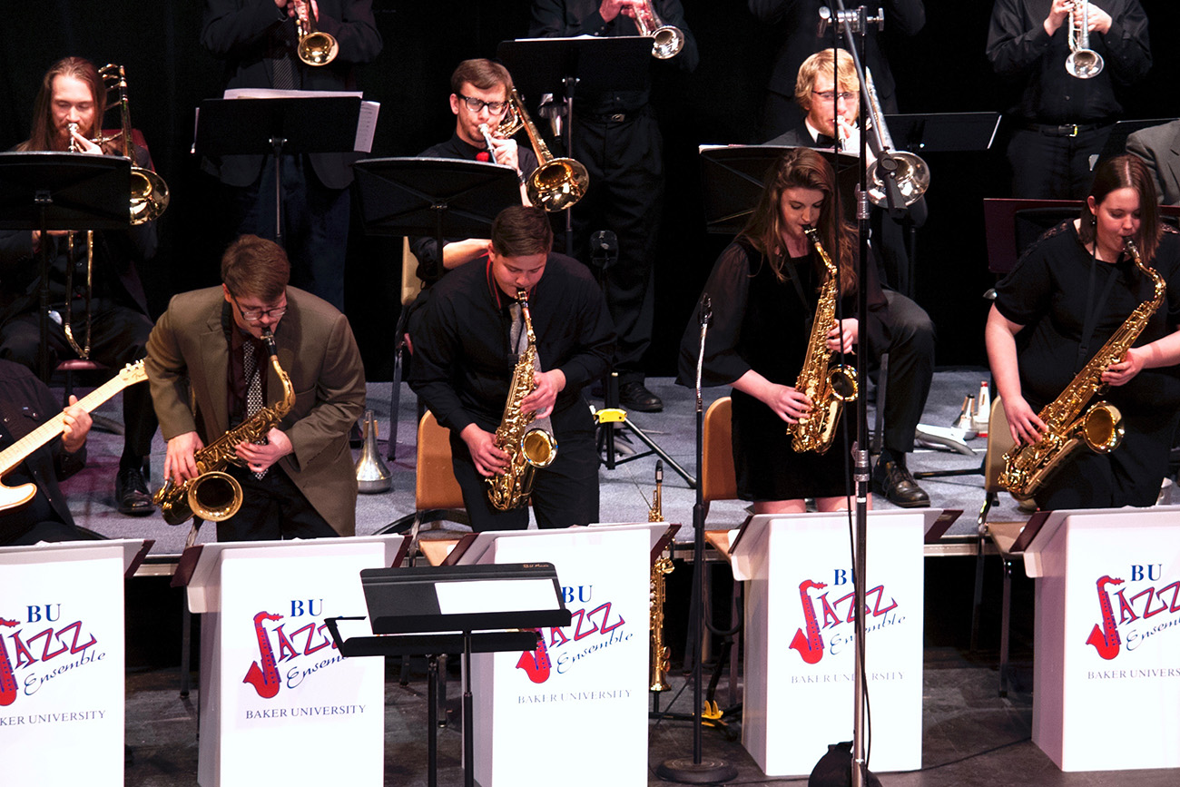 row of students playing the saxophone with a row of students playing the trombone behind them, at the jazz music festival