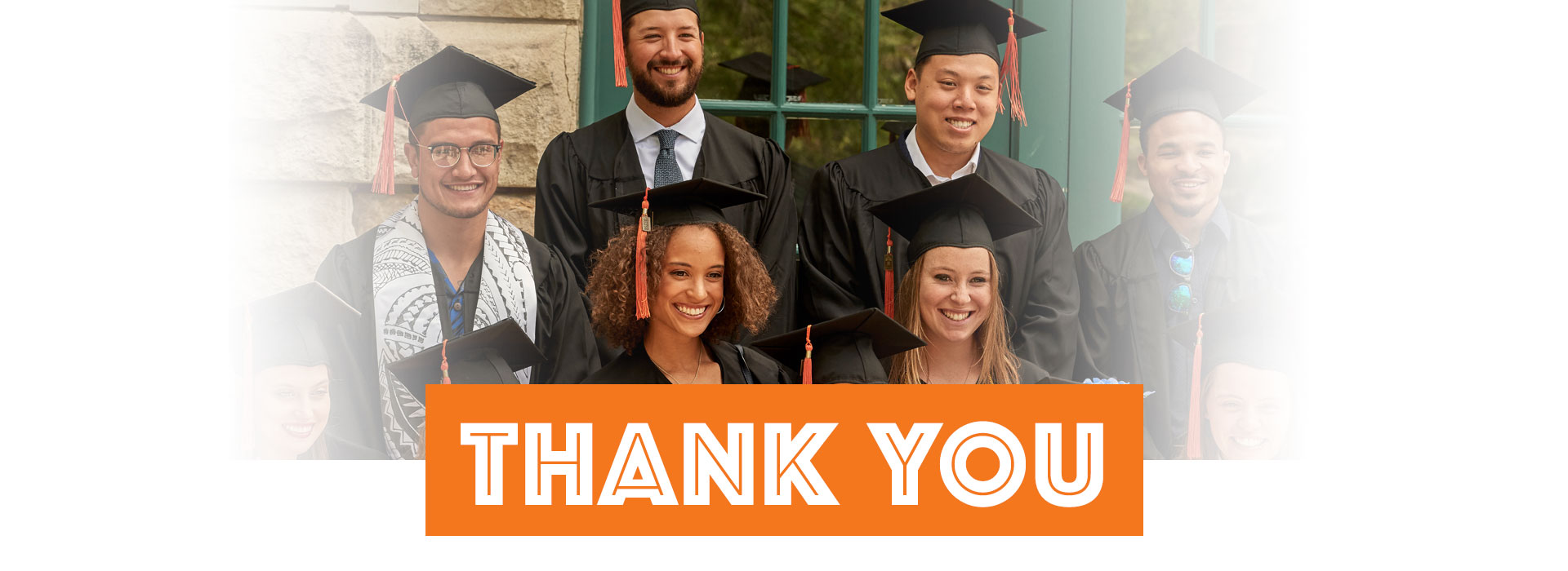 Graduate students in caps and gowns smiling with a thank you banner
