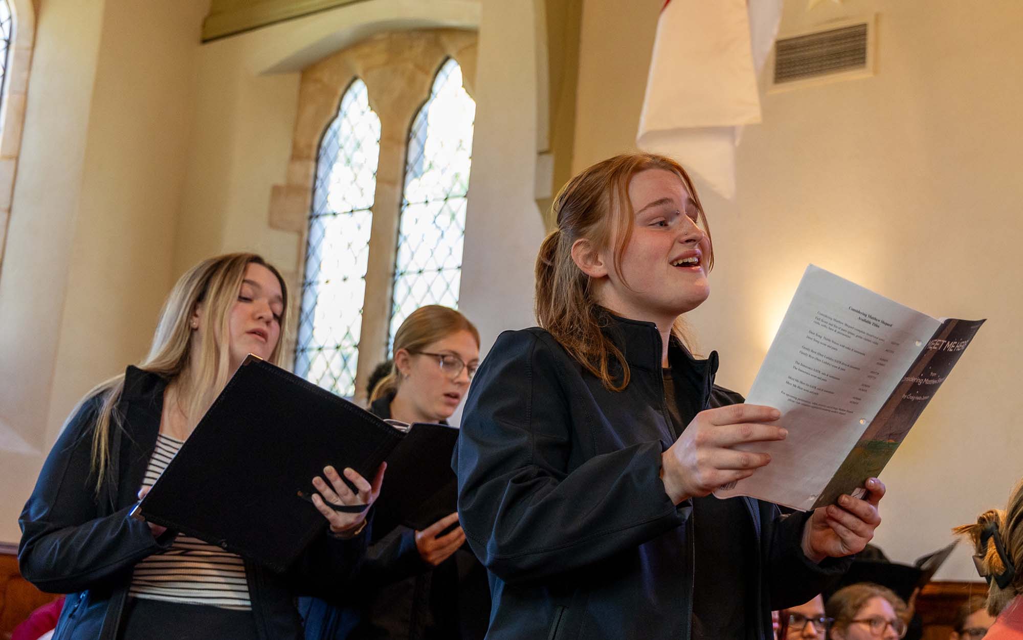 Three students holding music and singing from the pews in Osborne Chapel
